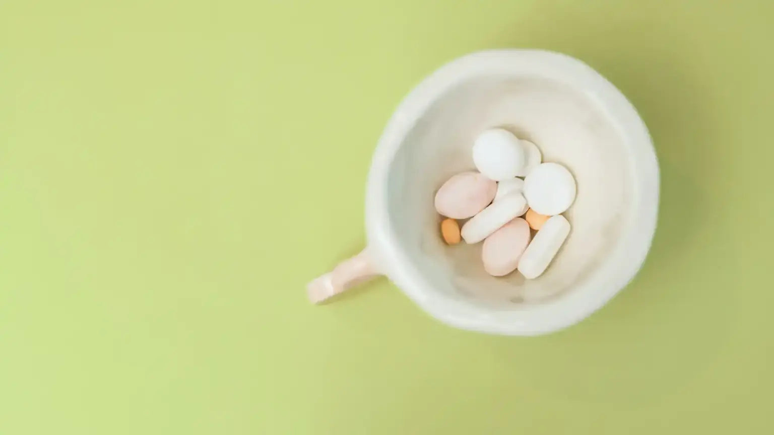 Top-down view of a white mug filled with assorted white, pink, and orange supplement tablets and capsules against a green background, illustrating NMN vs. NAD comparison of supplement forms and benefits.