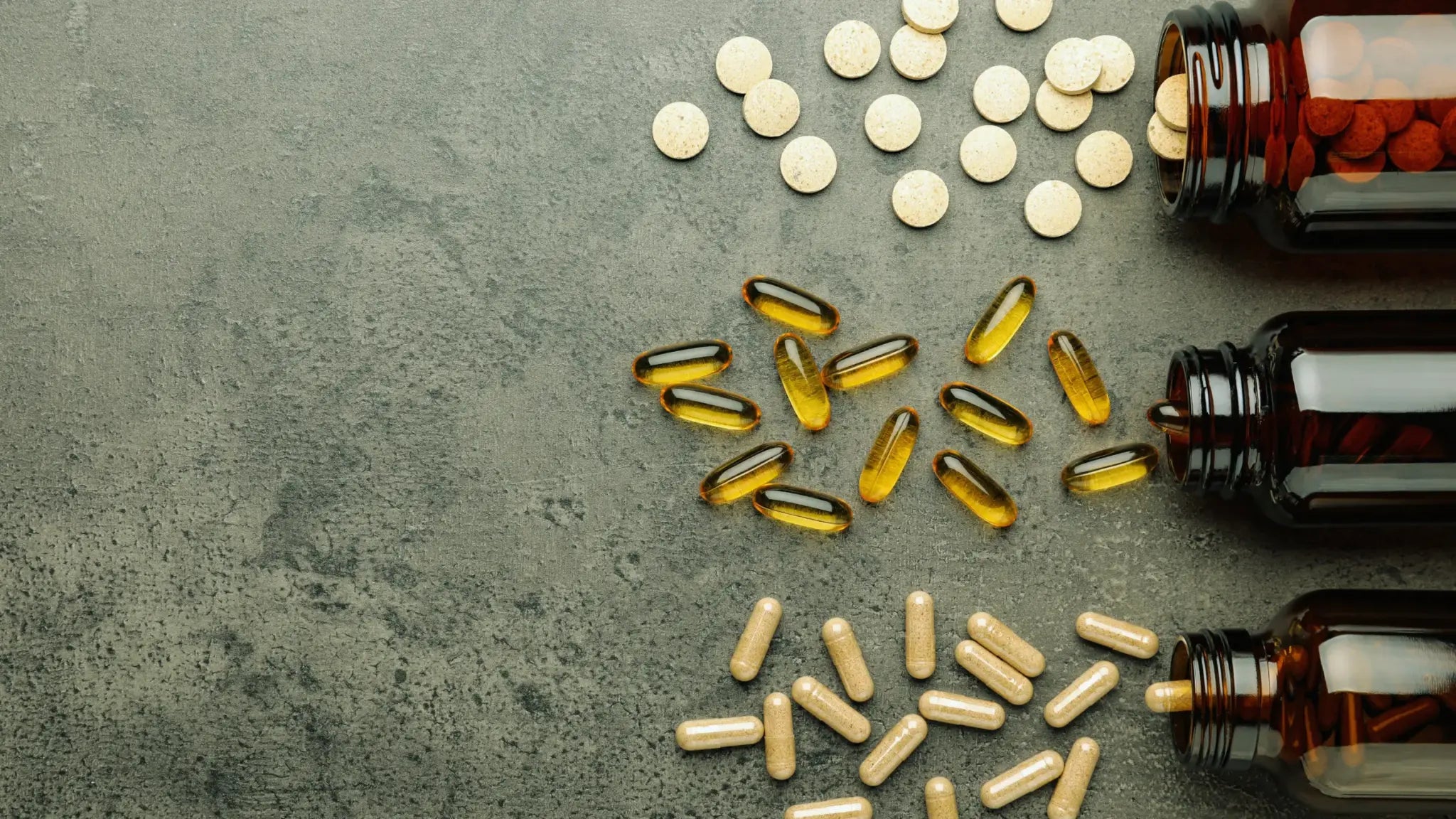 Top-down view of three amber supplement bottles tipped over on a gray stone surface, spilling yellow softgel capsules, beige round tablets, and beige capsules to illustrate Alpha-GPC vs. choline supplements.