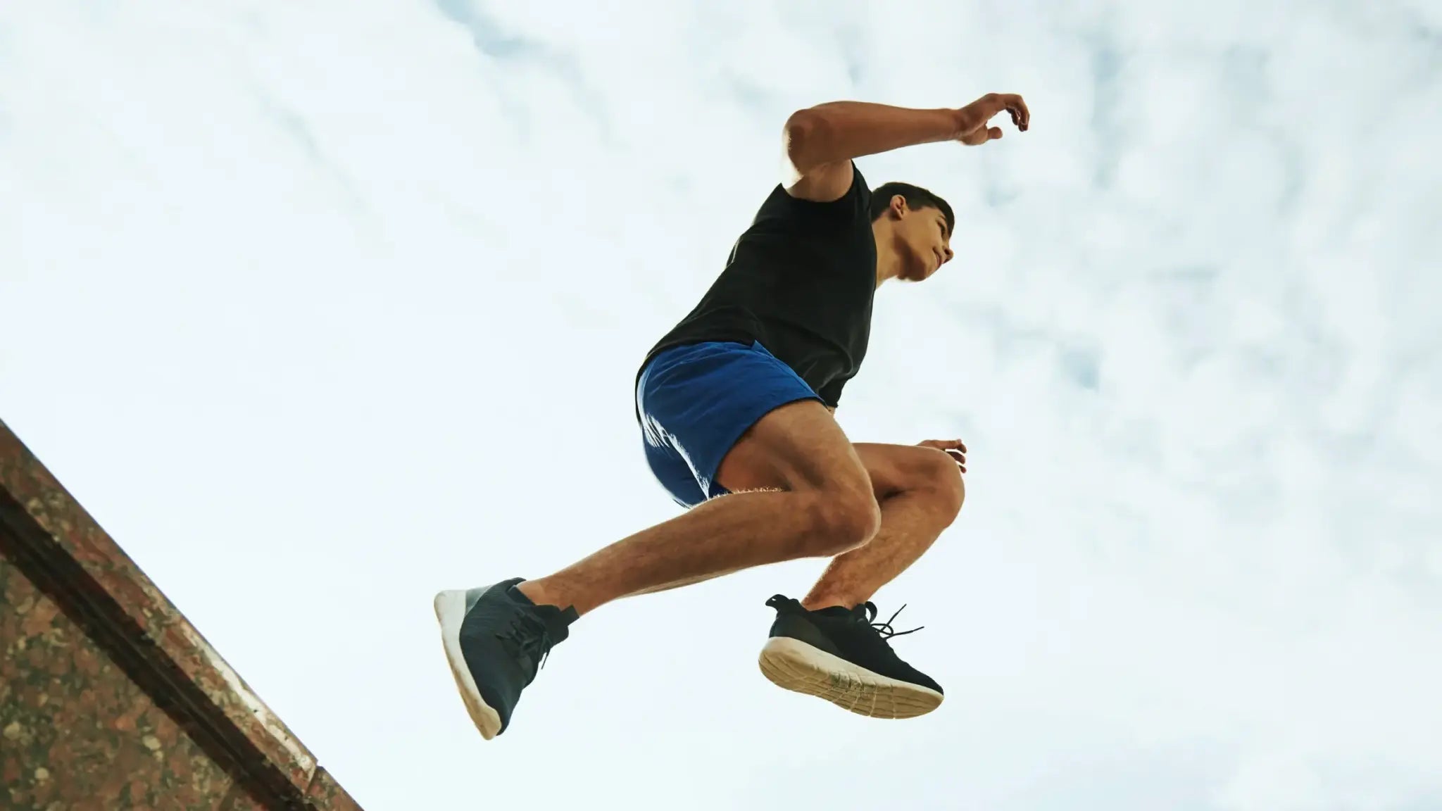 Low-angle shot of a young man in a black shirt and blue shorts leaping into the air against a bright sky, symbolizing the energetic, anti-aging benefits of NAD and resveratrol.