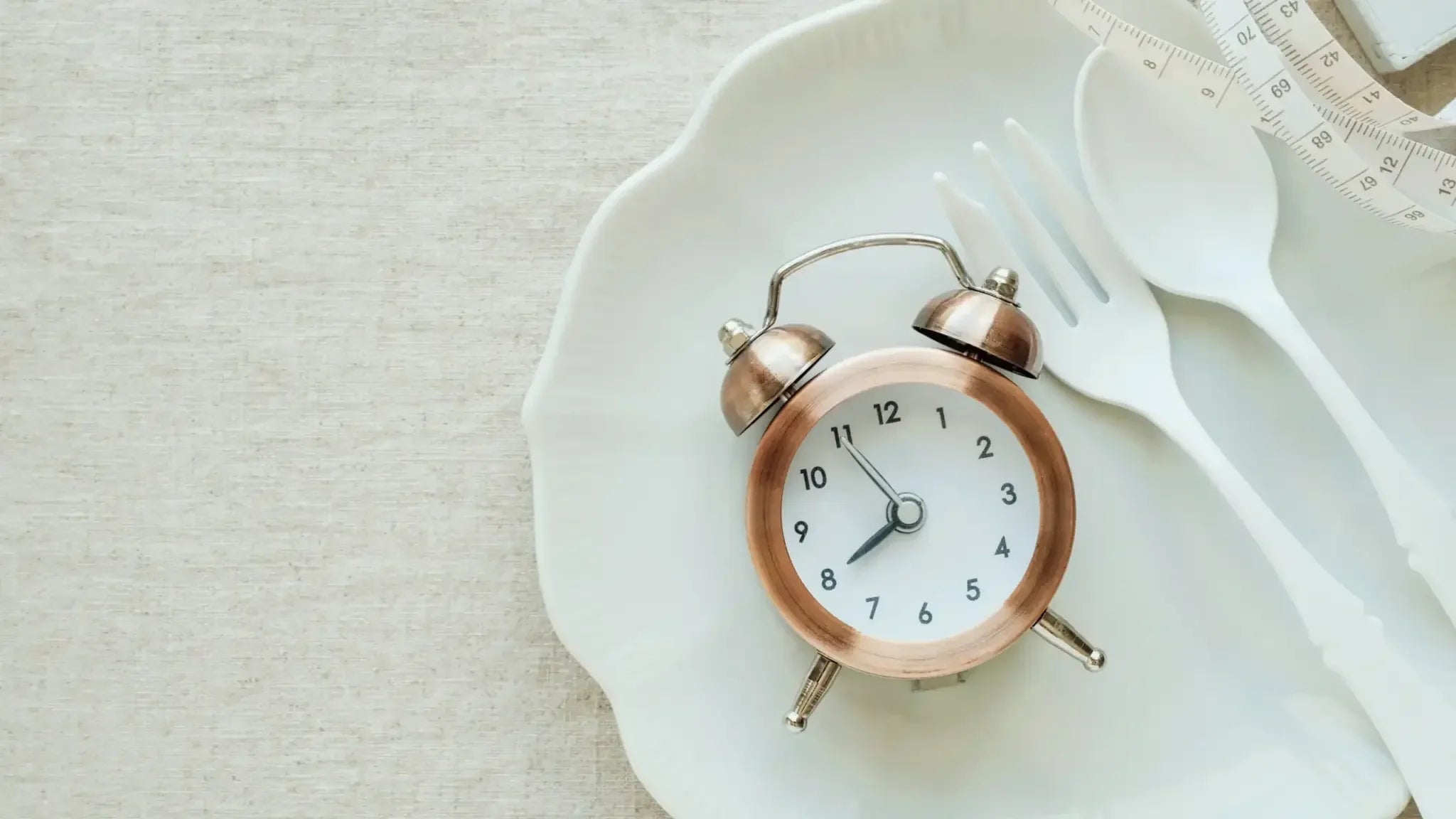 Copper alarm clock set to 10:10 resting on a white dinner plate with a fork, spoon, and measuring tape draped nearby, symbolizing intermittent fasting and its anti-aging benefits.
