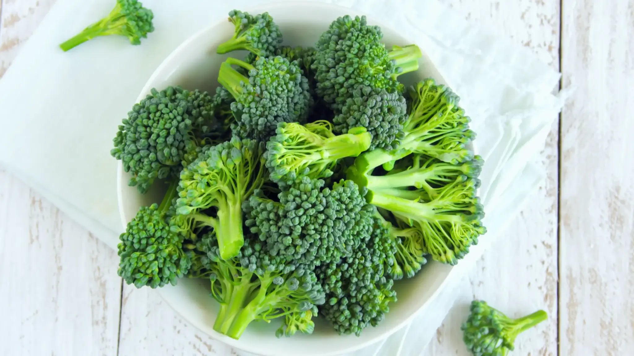 A white bowl filled with fresh green broccoli florets on a pale wooden surface, illustrating natural food sources of NMN.