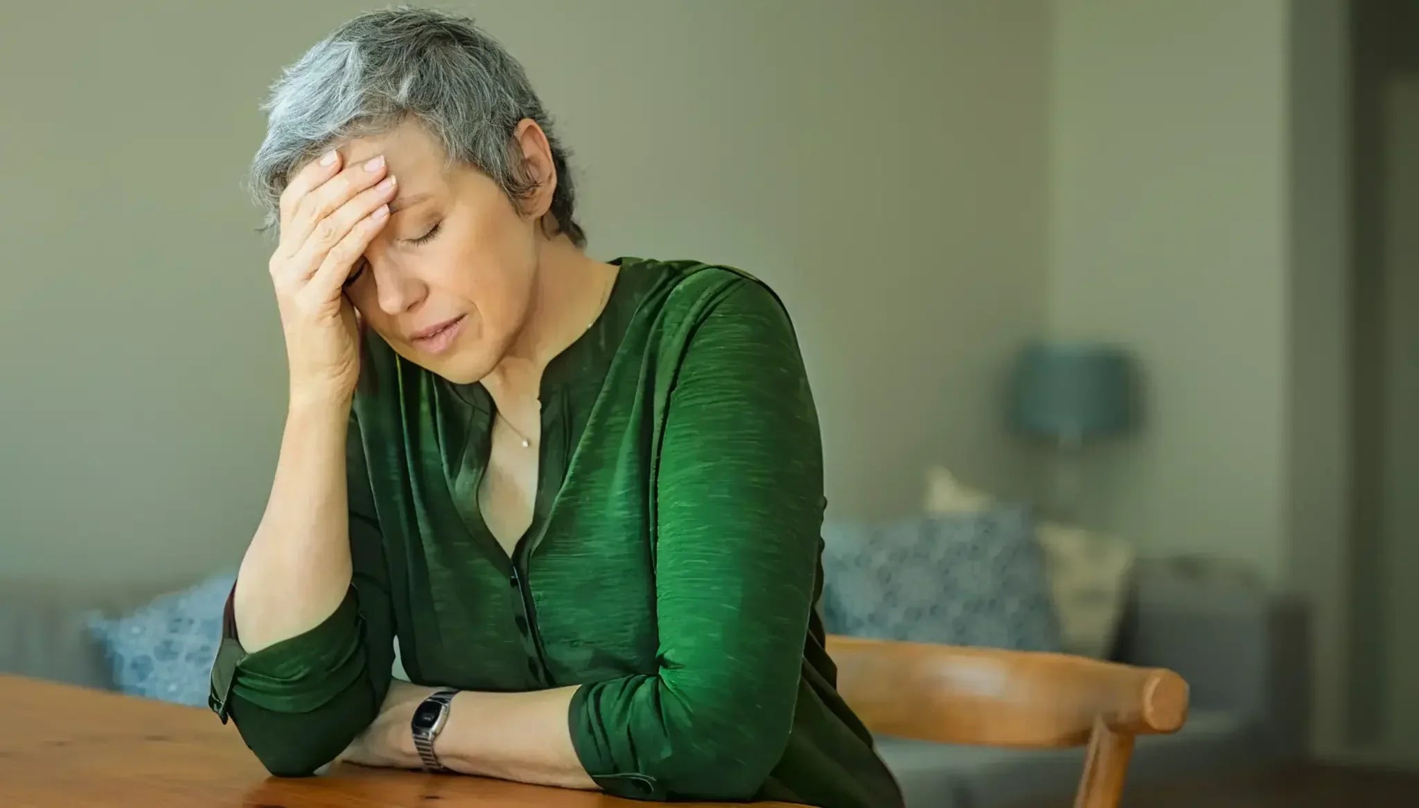 An older woman sits at a wooden table with her eyes closed and one hand pressed to her forehead in a gesture of fatigue and stress—illustrating the impact of chronic stress on health and lifespan and the need for effective stress-reduction strategies.
