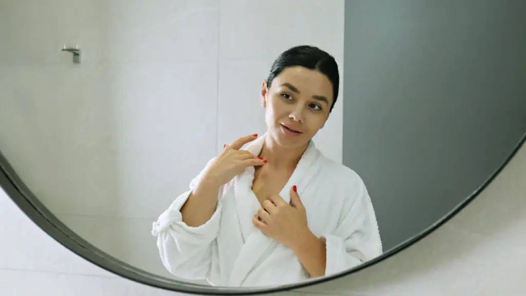 A woman wearing a white robe, looking at her reflection in a mirror