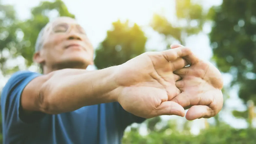 An elderly man stretches his arms while standing in a sunny park, surrounded by lush green trees