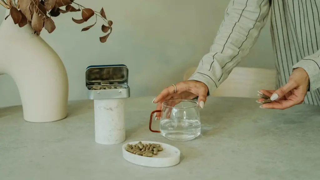 A woman holds a glass of water, preparing to take supplements from a bowl on the table