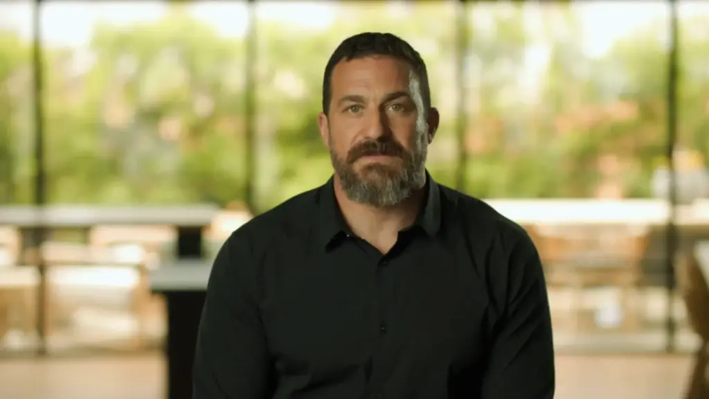 Andrew Huberman sitting on a chair, wearing a black T-shirt, with a focused expression